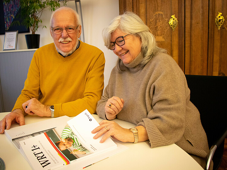 Ein Mann und eine Frau sitzen an einem Tisch, auf dem ein Buch mit der Aufschrift "WRT1+" liegt, in dem die beiden blättern.