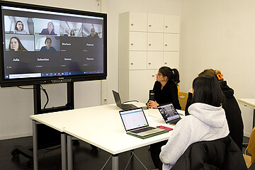 Drei Personen sitzen an einem Tisch vor Laptops, im Hintergrund ist ein großer Monitor zu sehen, an dem weitere Personen per Videokonferenz zugeschalten sind.