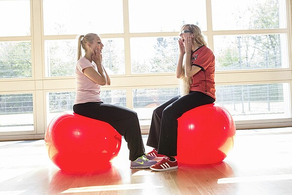 Auf dem Foto sind zwei Frauen auf roten Gymnastikbällen zu sehen. Beide halten ihre Hände an den Kiefer und haben den Mund weit geöffnet. Die beiden Frauen machen zusammen Sprachübungen.