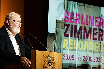 Das Bild zeigt einen Mann an einem Rednerpult, darauf ist das Wappen des Bundeslandes Baden-Württemberg zu erkennen. Im Hintergrund ist eine Projektion an der Wand zu sehen, darauf der Text "Berliner Zimmer Reloaded".