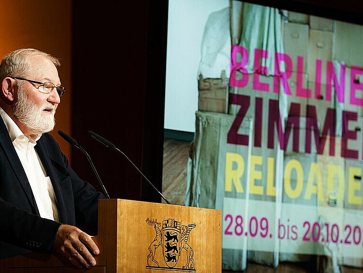Das Bild zeigt einen Mann an einem Rednerpult, darauf ist das Wappen des Bundeslandes Baden-Württemberg zu erkennen. Im Hintergrund ist eine Projektion an der Wand zu sehen, darauf der Text "Berliner Zimmer Reloaded".