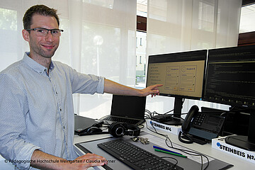 Simon Schaudt zeigt mit seiner Hand auf den PC-Monitor. Er steht vor seinem Arbeitsplatz in einem Büro der PH Weingarten.