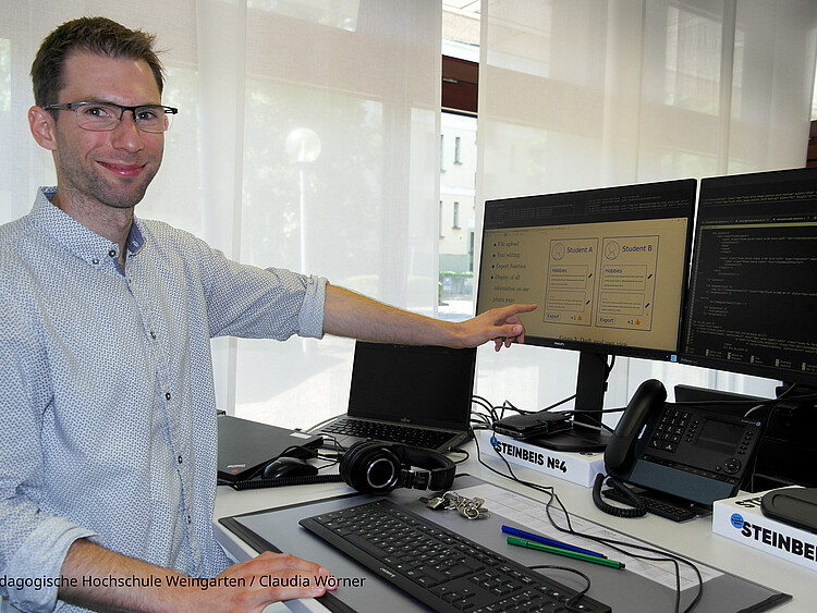 Simon Schaudt zeigt mit seiner Hand auf den PC-Monitor. Er steht vor seinem Arbeitsplatz in einem Büro der PH Weingarten.