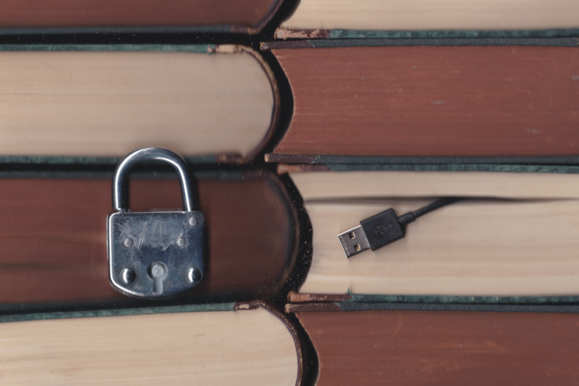 Ein Vorhängeschloss vor einem Bücherstapel sowie ein USB-Kabel, das aus einem der Bücher herausschaut. Datenschutz & Informationssicherheit